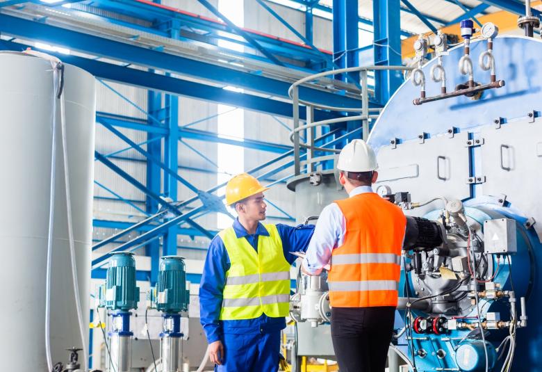 An employee and a manager in an industrial factory are talking to one another in the warehouse next to a large machine.