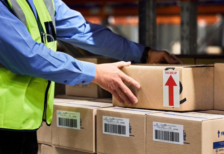 A man in a yellow safety vest is applying a shipping label to a cardboard box beside warehouse packing supplies.