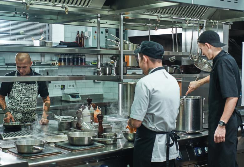 Three male workers wearing aprons cooking around a large stainless steel stove inside a commercial kitchen.