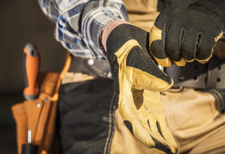 A close-up of a person wearing a plaid shirt and a tool belt putting on a pair of yellow work gloves.