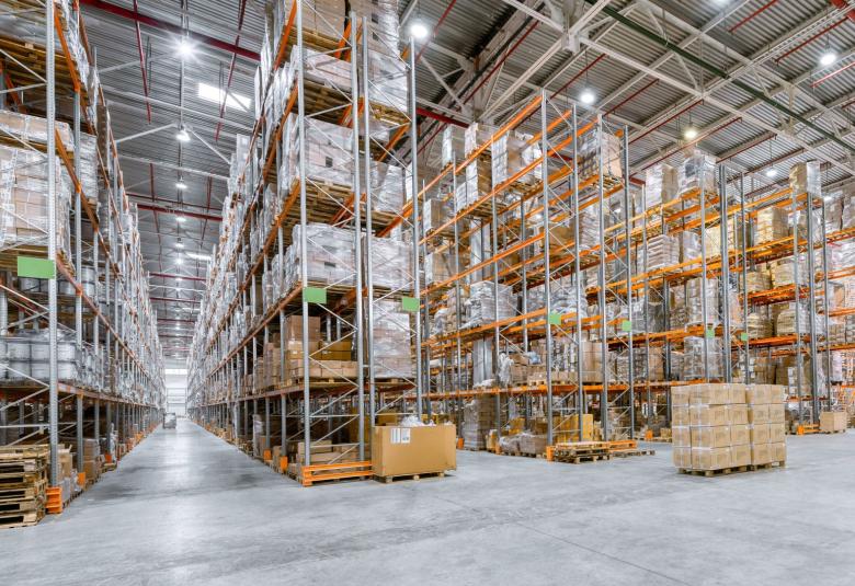 High bay lights are spaced evenly apart on a high warehouse ceiling. They evenly illuminate aisles and a large open space.
