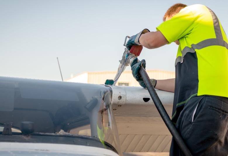 A mechanic fills up a plane's tank with fuel. The mechanic wears blue gloves and a yellow and black shirt.