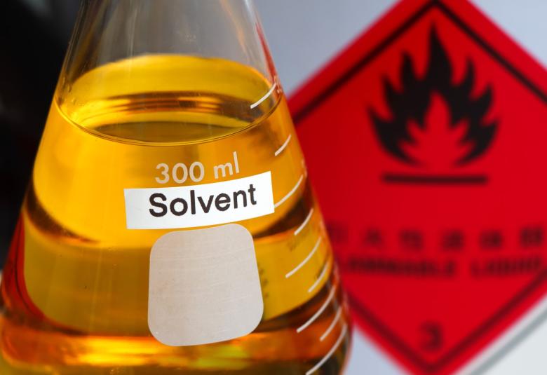 A beaker with a "solvent" label on the front is filled with orange liquid. There is a red flammable sign in the background.