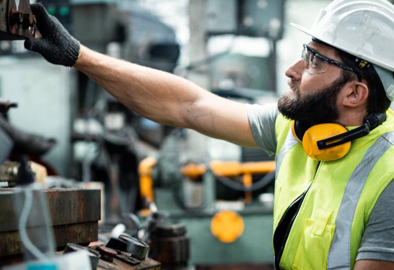 A man wearing a white helmet and other safety gear, working in an industrial warehouse, and checking up on the machinery.