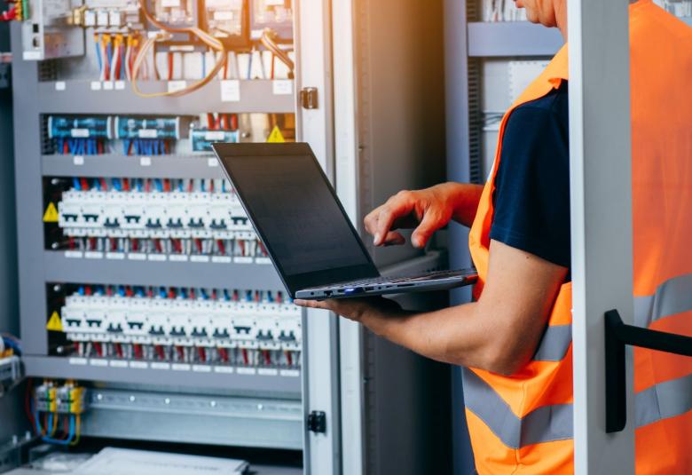 An electrician is repairing an electricity power station and checking the control cabinet with a laptop in his hands.