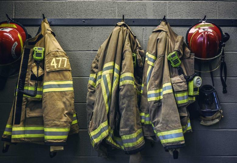 Three firefighter jackets hang alongside two red helmets on black hooks attached to a black cement wall.