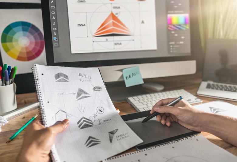 A close-up of a person sitting at a desk and working on a laptop that shows a logo. They hold a sketchbook in one hand with sketches of the same logo.