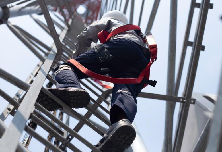 An engineer in a hard hat is climbing up a ladder on a communication tower while wearing a red safety harness.