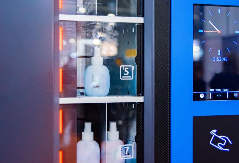 A high-tech industrial vending machine has bottles of antiseptic solution inside. There is a screen with an analog clock.